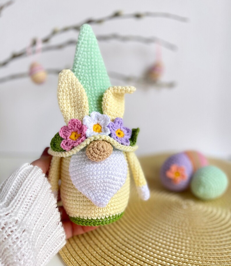 May include: A crocheted Easter gnome with a green hat, yellow body, white beard, and a flower crown. The gnome is holding a green and yellow Easter egg.