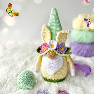 May include: A crocheted gnome with a yellow body, green hat, and white beard. The gnome is wearing a flower crown with pink, white, and yellow flowers. The gnome is sitting on a white fluffy surface with green and purple flowers.