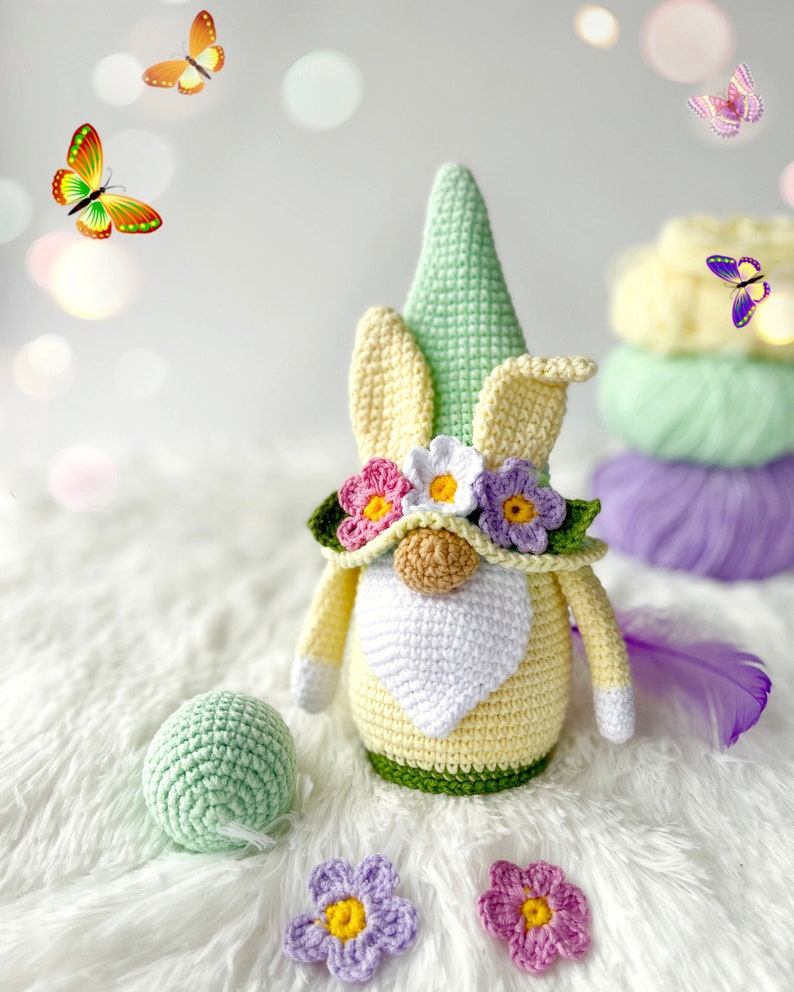 May include: A crocheted gnome with a yellow body, green hat, and white beard. The gnome is wearing a flower crown with pink, white, and yellow flowers. The gnome is sitting on a white fluffy surface with green and purple flowers.