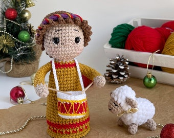 May include: A crocheted doll dressed in a yellow and red robe, holding a small drum. The doll has brown hair and a purple headband. A white crocheted lamb is standing next to the doll.