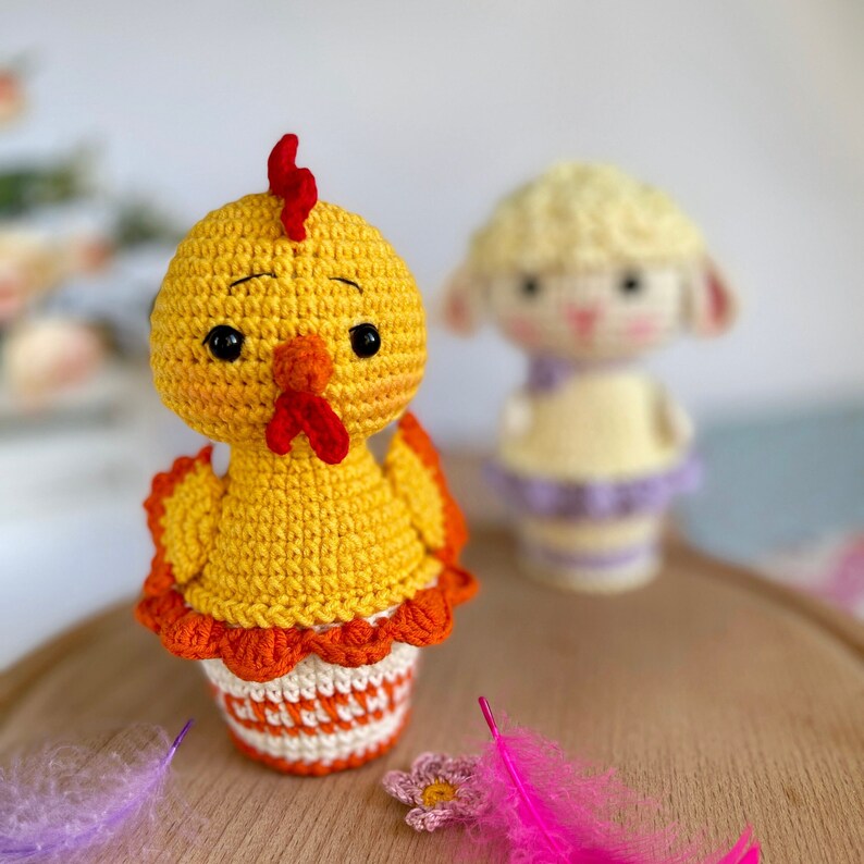 May include: A crocheted yellow chick with a red comb and orange ruffle sits in a white and orange crocheted pot. A crocheted white lamb with purple accents is in the background.