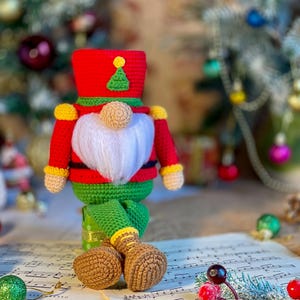 Puede incluir: Un juguete de gnomo de crochet vestido como un cascanueces, con un sombrero rojo con un árbol verde en la parte superior, una chaqueta verde y botas marrones. El gnomo está sentado en una hoja de música con una rama de bayas rojas y hojas verdes.