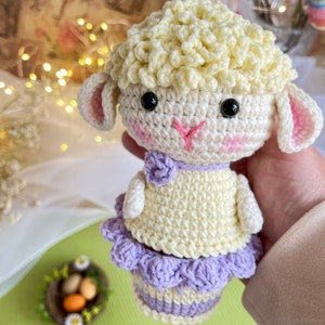May include: A crocheted Easter lamb with a white body, yellow curly hair, and a purple skirt. The lamb is holding a basket of eggs.