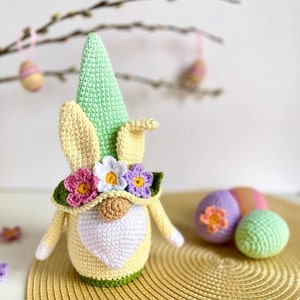 May include: A crocheted Easter gnome with a green hat, yellow bunny ears, and a flower crown. The gnome is holding a heart-shaped white beard and is standing on a gold placemat. There are two crocheted Easter eggs in the background.