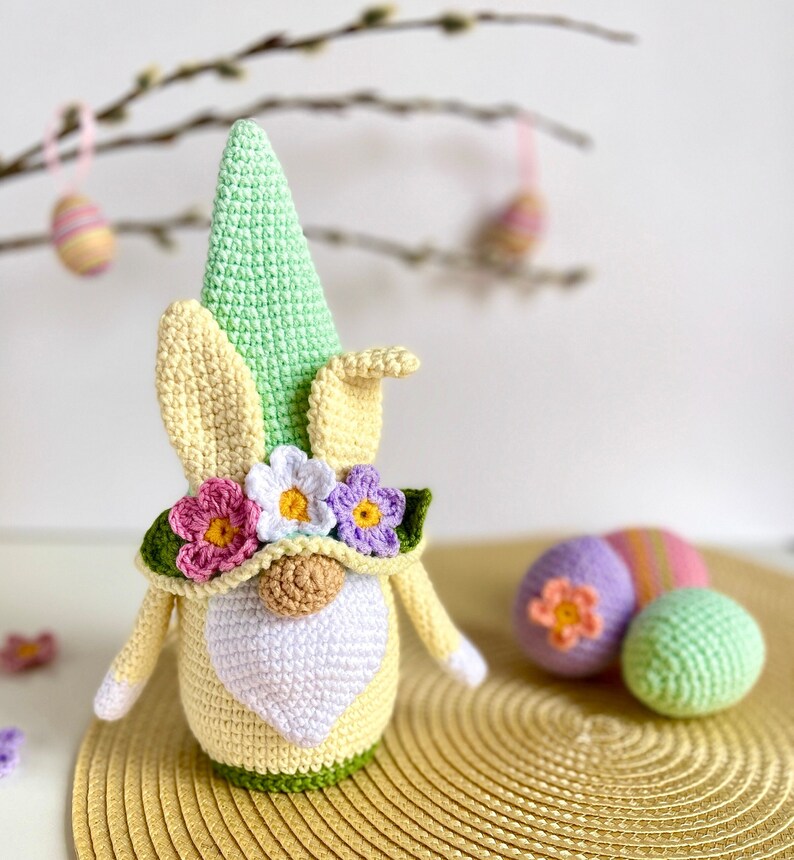 May include: A crocheted Easter gnome with a green hat, yellow bunny ears, and a flower crown. The gnome is holding a heart-shaped white beard and is standing on a gold placemat. There are two crocheted Easter eggs in the background.