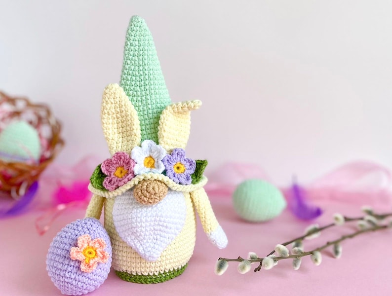 May include: A crocheted Easter gnome with a green hat, yellow ears, and a white beard. The gnome is wearing a flower crown and holding a purple Easter egg.