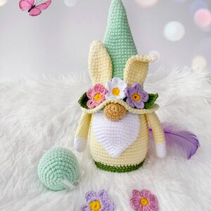 May include: A crocheted gnome with a green hat, yellow and white body, and a flower crown. The gnome is sitting on a white fluffy surface with a green ball and two crocheted flowers.
