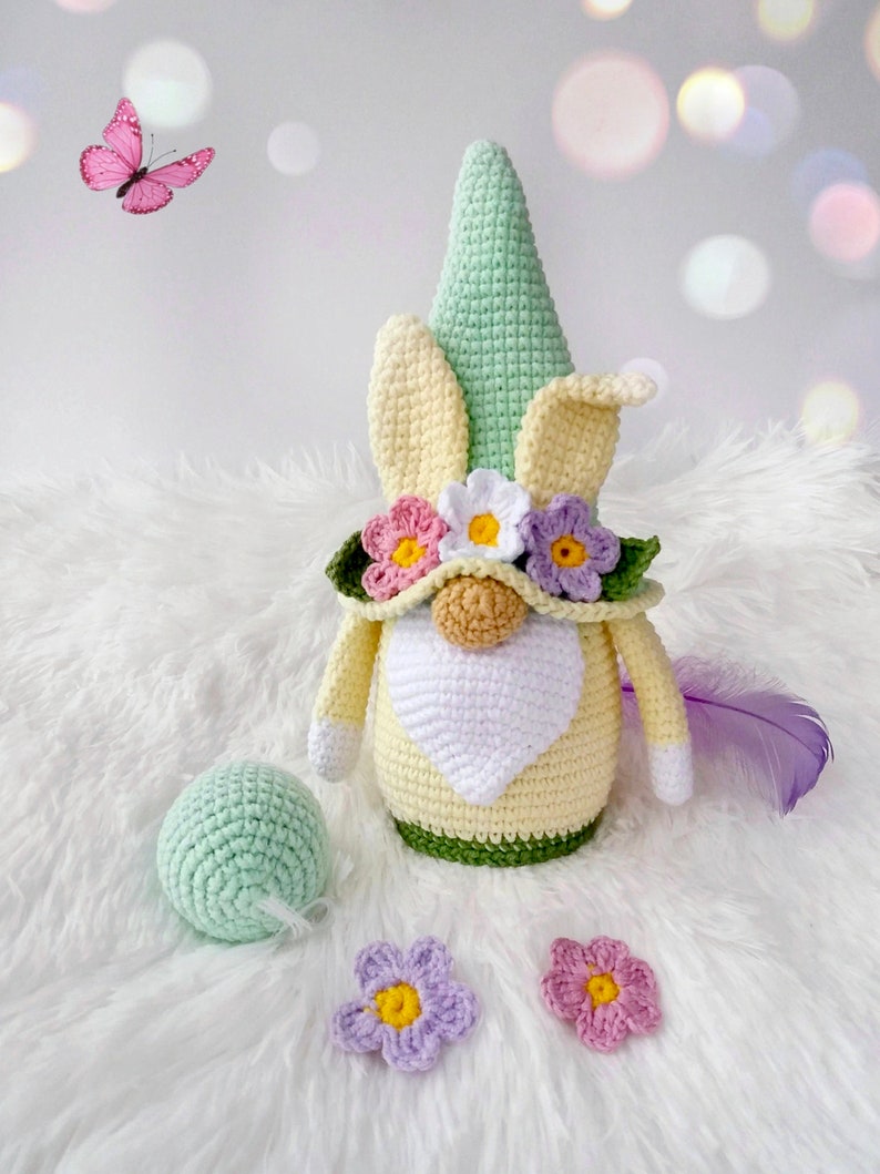 May include: A crocheted gnome with a green hat, yellow and white body, and a flower crown. The gnome is sitting on a white fluffy surface with a green ball and two crocheted flowers.