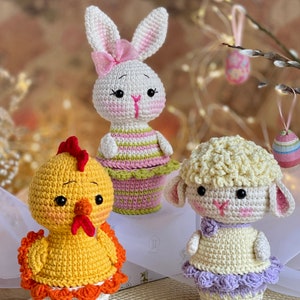 Può includere: Tre animali all'uncinetto pasquali in vasi a forma di cupcake colorati. Un coniglietto bianco con orecchie rosa e un fiocco, un pulcino giallo con un becco arancione e un agnello bianco con una gonna viola.