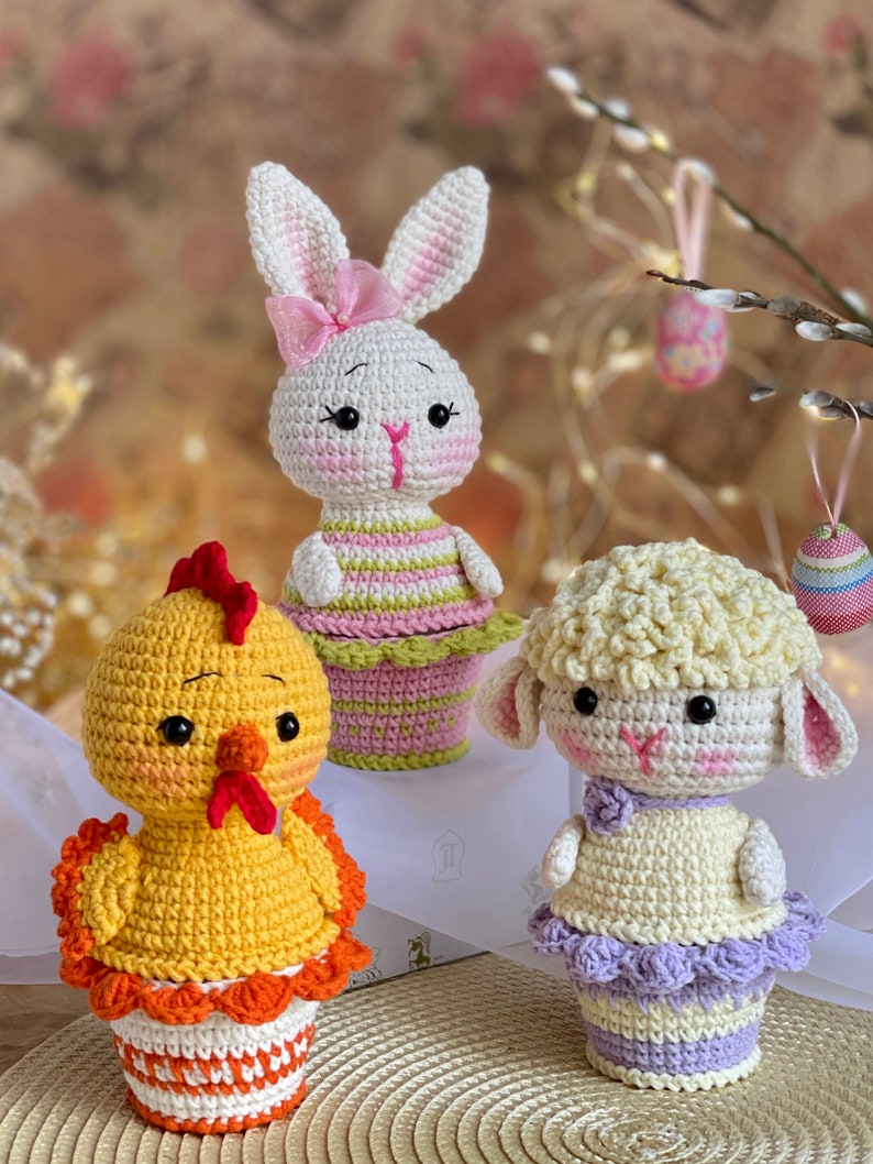 May include: Three crocheted Easter animals in colorful cupcake-shaped pots. A white bunny with pink ears and a bow, a yellow chick with an orange beak, and a white lamb with a purple skirt.