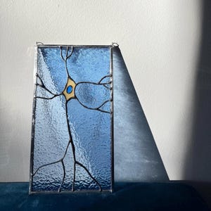 May include: A rectangular stained glass panel featuring a blue textured glass background with a central cream-colored neuron shape. The neuron has a small blue circular center and dark metal solder lines forming branching dendrites and an axon.