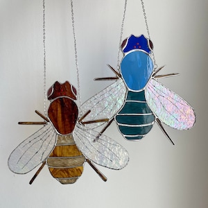 以下が含まれることがあります： 銀色のチェーンで吊るされた、2つのステンドグラスのハエの飾り。1つは茶色の頭と黄色の体、もう1つは青い頭とティール色の体です。どちらも虹色の羽があり、白い背景に配置されています。