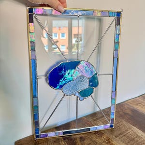 Può includere: Un pannello in vetro colorato rettangolare con un design centrale a forma di cervello composto da varie tonalità di blu, verde acqua e vetro iridescente. Il cervello è incorniciato da un bordo di piccoli quadrati di vetro multicolore e sottili linee di saldatura.