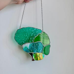 Könnte beinhalten: Ein Buntglas-Suncatcher in Form eines menschlichen Gehirns, zusammengesetzt aus verschiedenen grünen und türkisfarbenen Glasstücken, die durch silberne Lötlinien verbunden sind. Das Objekt hängt an einer dünnen silbernen Metallkette.