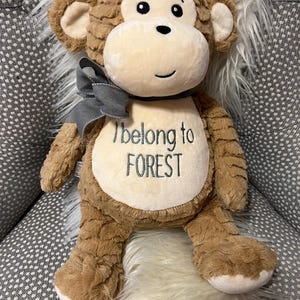May include: A plush monkey with brown fur, a cream-colored face, and a gray bow. The text "I belong to FOREST" is embroidered on the monkey's chest. The monkey is sitting on a chair with a white fur pillow.