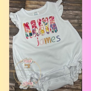 May include: A white baby onesie with a colorful floral fabric applique spelling out the name "Maya James".