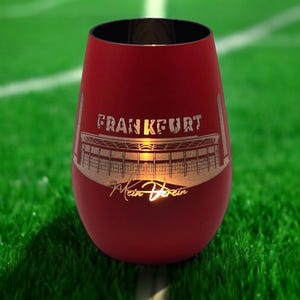 Op de afbeelding: Een rode wijnglas zonder voet met het woord "FRANKFURT" en een stadionontwerp in goud. Het glas heeft een zwarte rand en staat op groen gras. De tekst "Mein Verein" is ook zichtbaar.