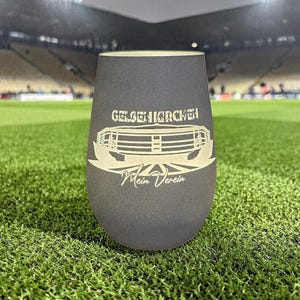 Könnte beinhalten: Ein dunkelgraues Weinglas ohne Stiel mit den weißen Schriftzügen "GELSENKIRCHEN" und "Mein Verein" sowie einer Stadiongrafik. Das Glas steht auf einem grünen Rasenfeld mit einem Stadion im Hintergrund.