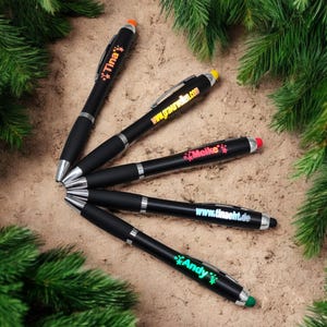 Könnte beinhalten: Fünf schwarze Stifte mit silbernen Akzenten und farbigen Touch-Spitzen liegen auf einer sandigen Oberfläche, umgeben von grünem Laub. Jeder Stift trägt einen anderen Namen oder eine Website in einer leuchtenden Farbe, darunter "Tina" und "Meike".
