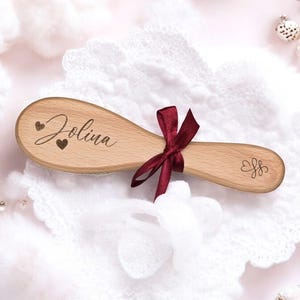 Könnte beinhalten: Eine Holzbürste für Babyhaar mit dem eingravierten Namen "Jolena". Die Bürste ist mit einem roten Band und zwei kleinen Herzen verziert.