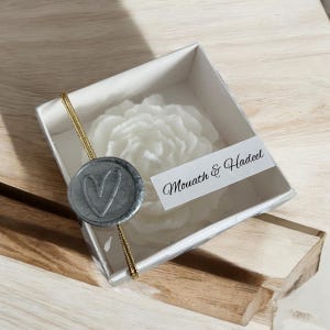 May include: A white wax flower candle in a clear box with a grey wax seal featuring a heart design. The box has a gold ribbon and a white label with the text "Mouath & Hadeel".