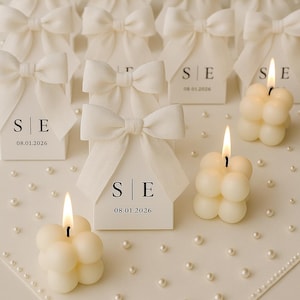 May include: Ivory-colored bubble candles with lit flames and small white gift boxes with bow accents. The boxes have the initials "S|E" and the date "08.01.2026" printed on them. The scene is set on a white surface with scattered pearls.