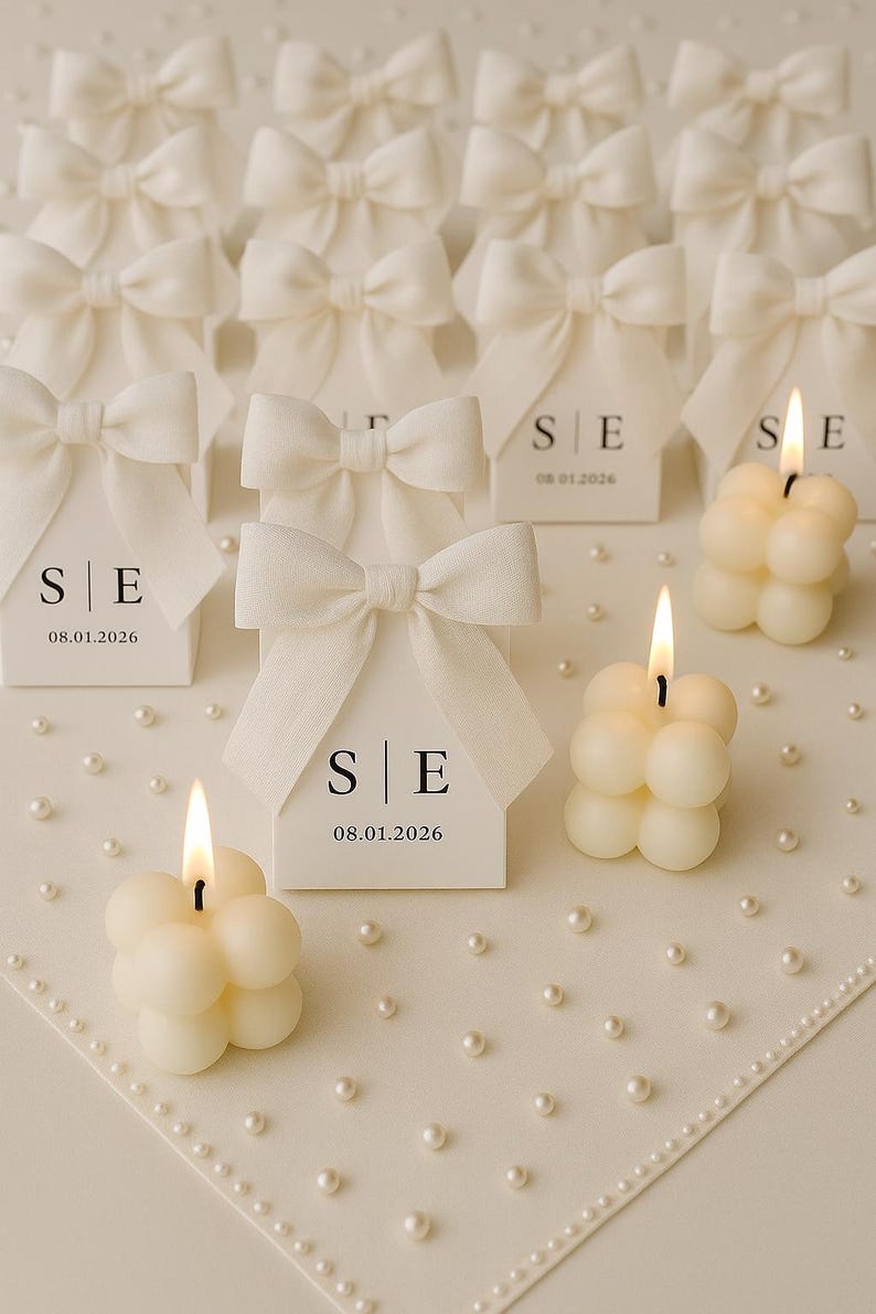 May include: Ivory-colored bubble candles with lit flames and small white gift boxes with bow accents. The boxes have the initials "S|E" and the date "08.01.2026" printed on them. The scene is set on a white surface with scattered pearls.