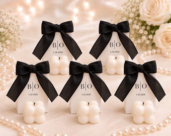 May include: Five white rectangular favor boxes each topped with a black satin ribbon bow. The front of each box features the black printed letters B|O and the date 1.10.2026. A small, off-white, square-shaped bubble candle sits in front of each box.