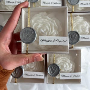 May include: White rose-shaped candles in small white boxes with a silver heart seal and the text "Mouath & Hadeel" on a white label.