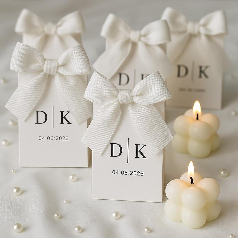 May include: White favor boxes with DK initials and the date 04.06.2026, tied with white bows. Two bubble candles are lit, and scattered pearls add a decorative touch. The boxes are likely for a wedding or special event.
