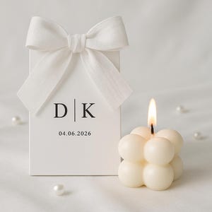 May include: A white gift box with a white bow, the initials "D | K", and the date "04.06.2026" printed on the front. A lit, ivory-colored bubble candle is next to the box. Several white pearls are scattered around.