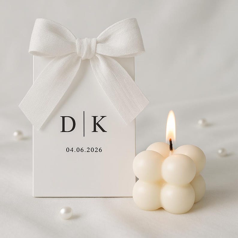 May include: A white gift box with a white bow, the initials "D | K", and the date "04.06.2026" printed on the front. A lit, ivory-colored bubble candle is next to the box. Several white pearls are scattered around.