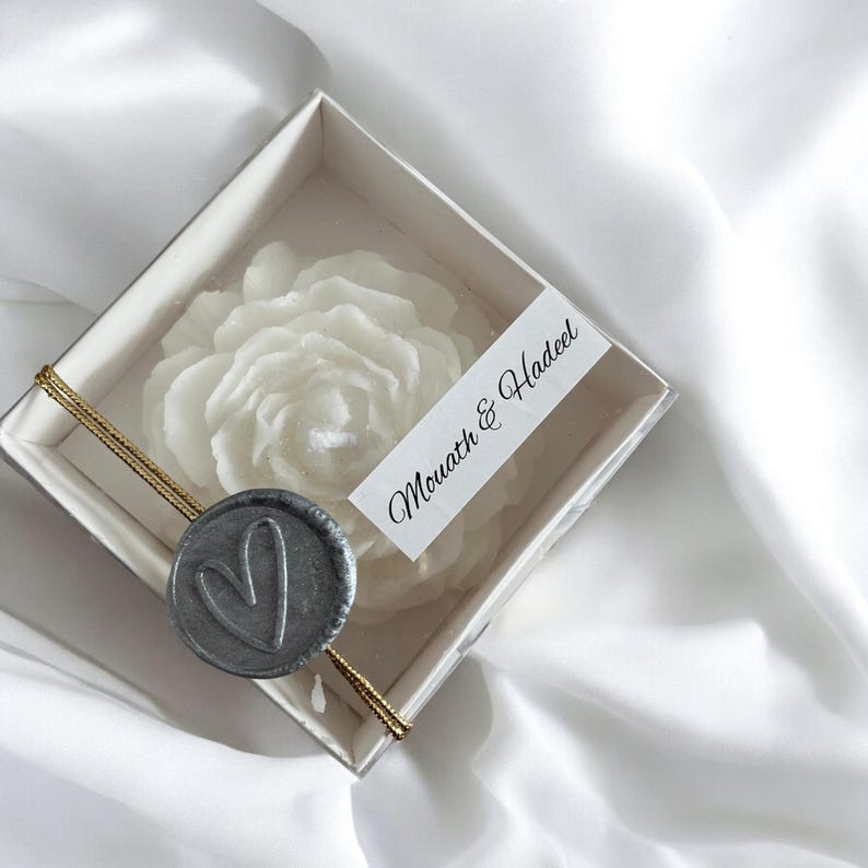 May include: A white rose-shaped candle in a white box with a gold ribbon and a grey wax seal with a heart design. The box has a white label with the text "Mouath & Hadeel".