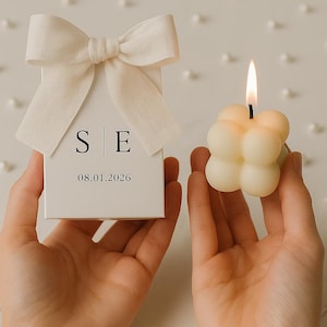 May include: A white gift box with a cream-colored bow, the initials "S E" and the date "08.01.2026" printed on the front. A lit, cream-colored bubble candle is held next to the box. The candle has a small, black wick.