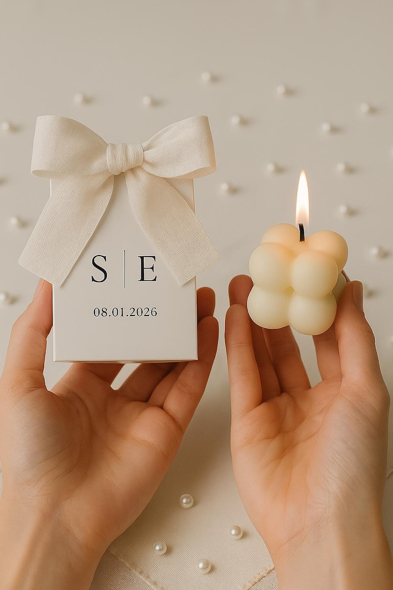 May include: A white gift box with a cream-colored bow, the initials "S E" and the date "08.01.2026" printed on the front. A lit, cream-colored bubble candle is held next to the box. The candle has a small, black wick.
