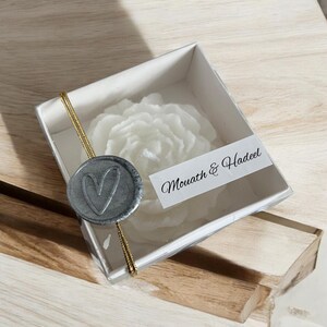 May include: A white wax flower candle in a clear box with a grey wax seal featuring a heart design. The box has a gold ribbon and a white label with the text "Mouath & Hadeel".