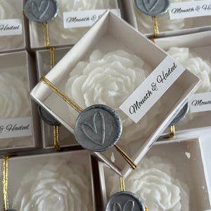 May include: White rose-shaped candles in clear boxes with gold ribbon and silver wax seals. The boxes have the text "Mouath & Hadeel" printed on them.