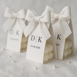 May include: Three white favor boxes with bubble-shaped candles inside, tied with white bows. Each box has a tag with initials and a date printed in black. Scattered pearls add a touch of elegance.