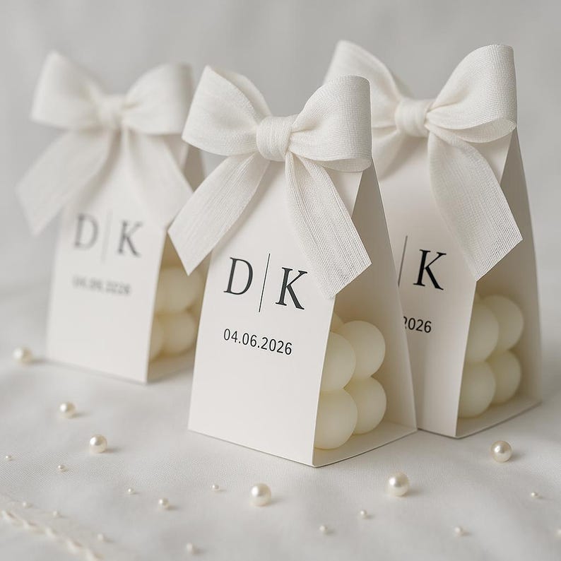 May include: Three white favor boxes with bubble-shaped candles inside, tied with white bows. Each box has a tag with initials and a date printed in black. Scattered pearls add a touch of elegance.