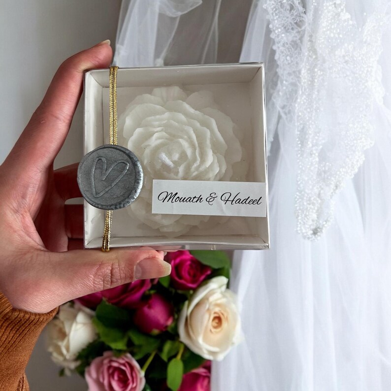 May include: A white rose-shaped candle in a clear box with a silver heart seal and a white label that reads "Mouath & Hadeel".