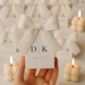 May include: A hand holds a white favor box with a large bow. The box displays the initials "D | K" and the date "04.06.2026". Several similar boxes and lit bubble candles are in the background, suggesting a wedding or event.