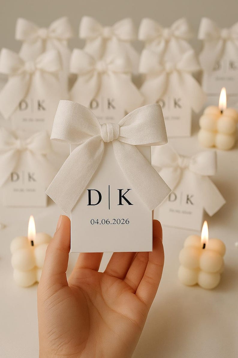 May include: A hand holds a white favor box with a large bow. The box displays the initials "D | K" and the date "04.06.2026". Several similar boxes and lit bubble candles are in the background, suggesting a wedding or event.