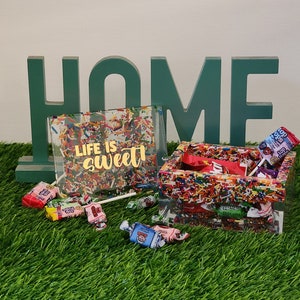 May include: A clear box with a colorful sprinkle design and the words "Life is sweet" in gold lettering. The box is filled with colorful candies and is sitting on a green artificial grass surface. The word "HOME" is spelled out in green letters behind the box.