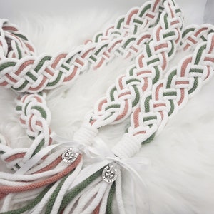 May include: Two white, green, and pink braided cords with tassels and silver charms. The cords are tied together with a white ribbon.