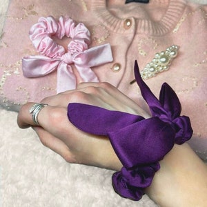 May include: A purple satin scrunchie tied in a bow with a pink satin scrunchie and bow on a pink cardigan with pearl embellishments.