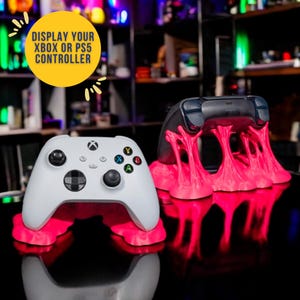 May include: Two video game controllers, one white and one black, are displayed on pink, abstract, 3D printed stands. The white controller is an Xbox controller, and the black controller is a Playstation controller. The text "DISPLAY YOUR XBOX OR PS5 CONTROLLER" is in a yellow circle above the controllers.