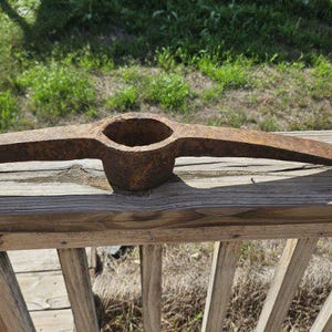 May include: A rusty, vintage pickaxe with a central eye and two curved blades. The tool is made of metal and rests on a weathered wooden surface, with a grassy background.