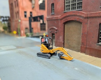 May include: A miniature yellow excavator model featuring black tracks, a black operator cabin with a small red light on top, and a movable articulated arm with a bucket, positioned on a grey surface in front of a red brick building model.