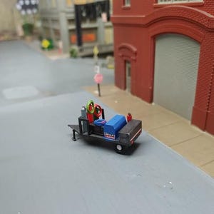 May include: A small, black welding cart with a blue and red welding machine, two green and yellow gas tanks, and a black hose. The cart has a black metal frame and a black metal platform. The cart is on a gray concrete surface.