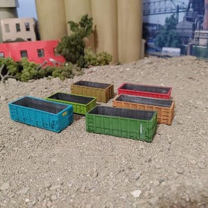 May include: Six miniature dumpsters in various colors, including blue, green, yellow, orange, and brown. The dumpsters are arranged in a row on a sandy surface.